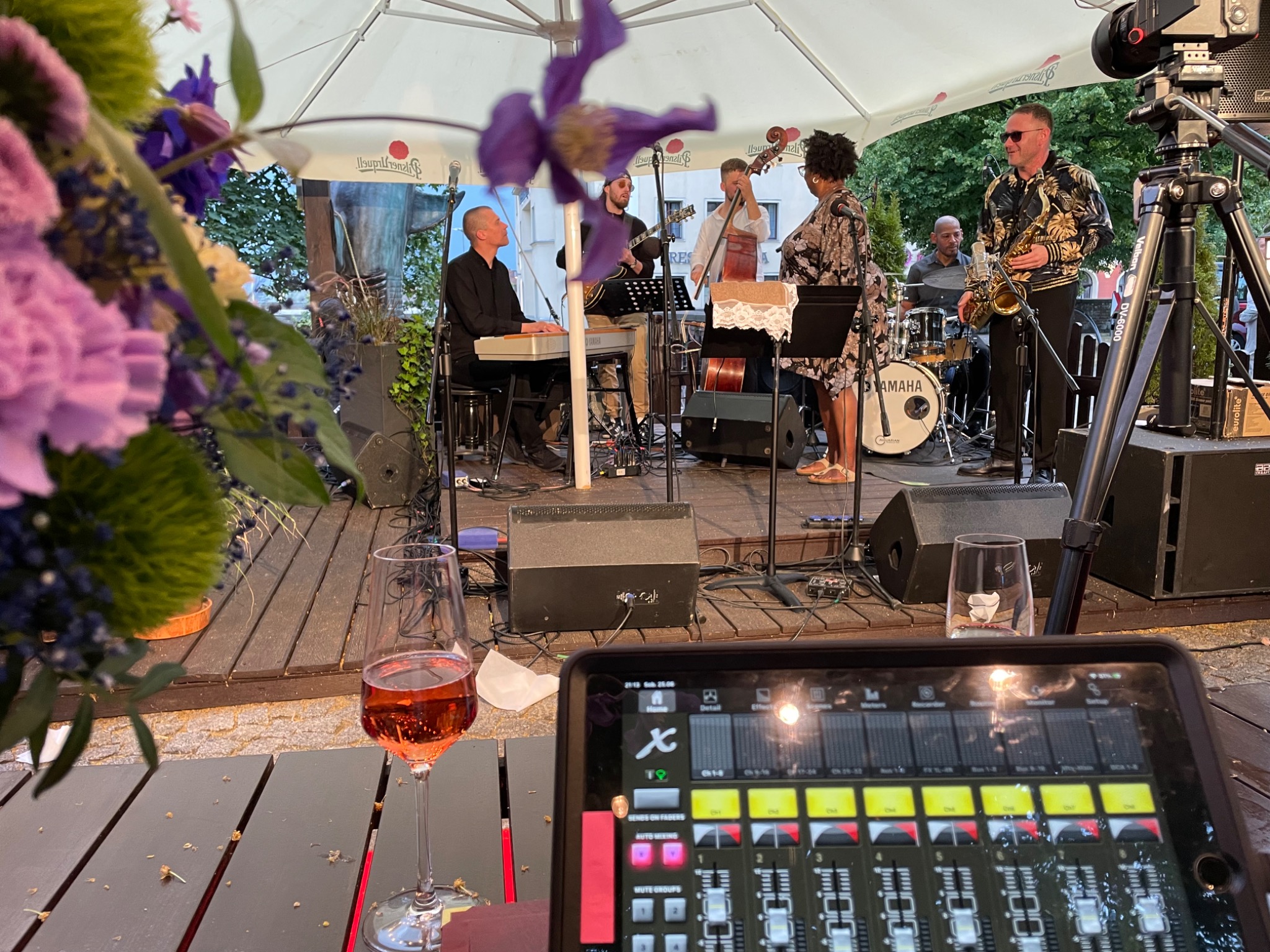 Zespół muzyczny na scenie plenerowej, widok zza stolika z kieliszkiem różowego wina i tabletem z interfejsem miksera dźwięku.