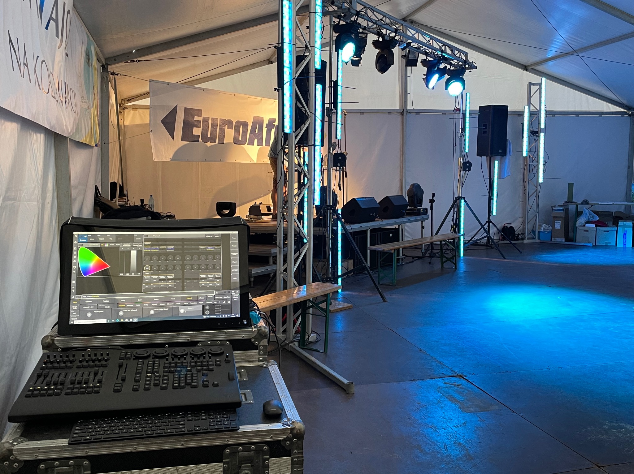 Stanowisko sterowania oświetleniem i nagłośnieniem w namiocie, widoczny panel sterowania z monitorem, reflektory na kratownicy oraz kolumny świetlne, podłoga oświetlona na niebiesko.