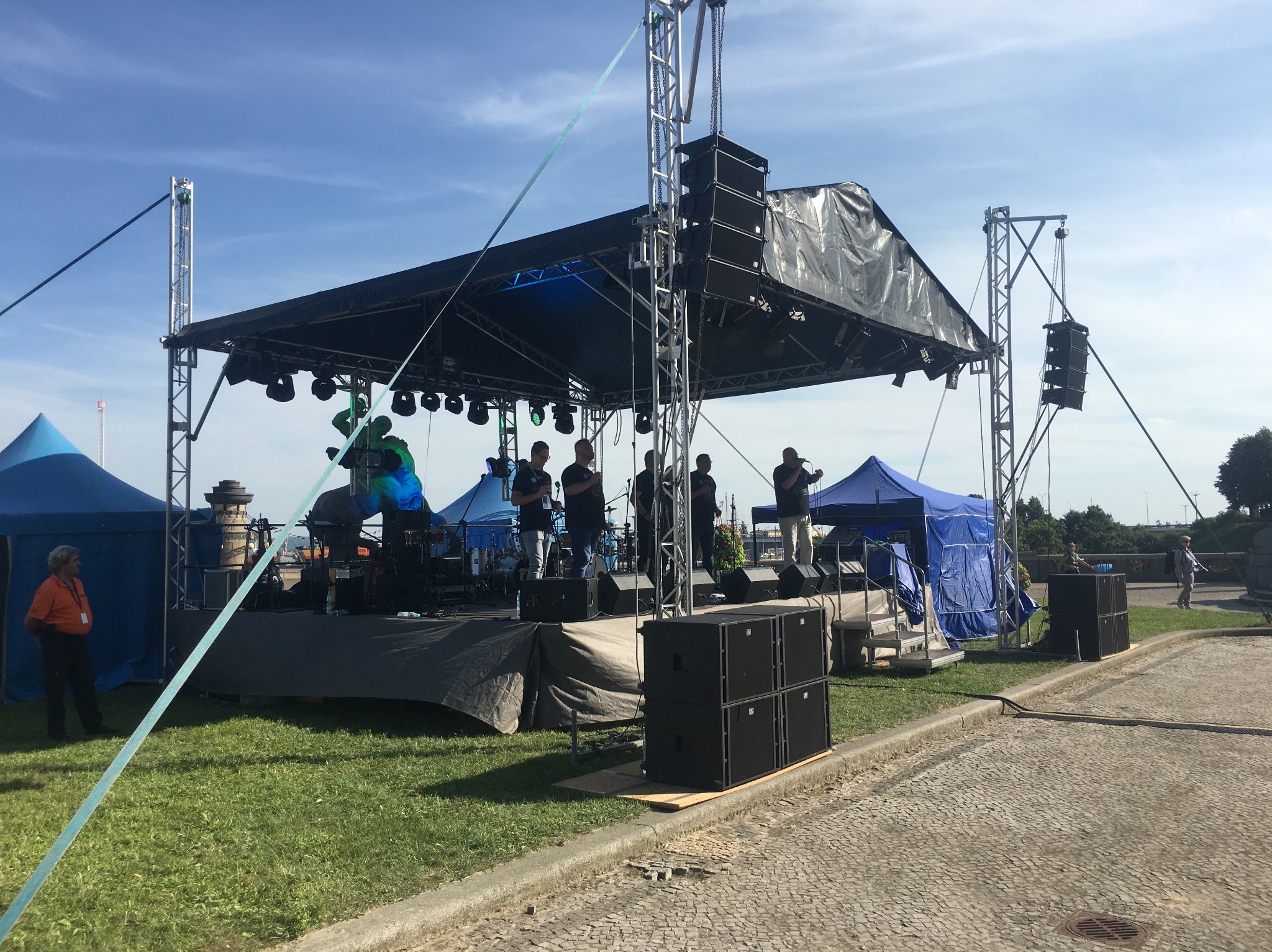 Scena plenerowa z zespołem podczas występu, widoczne kolumny głośnikowe, reflektory i zadaszenie, a także osoby obsługujące wydarzenie i publiczność w tle.