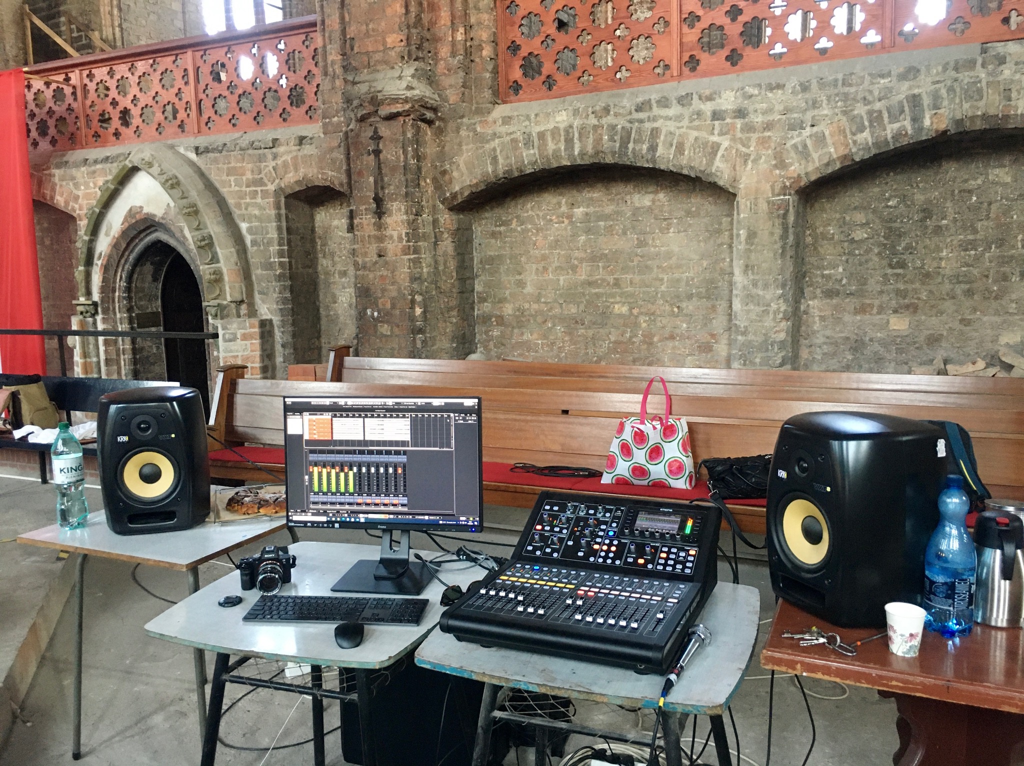 Stanowisko pracy z mikserem cyfrowym, monitorem z oprogramowaniem DAW, głośnikami studyjnymi KRK, aparatem fotograficznym Sony i butelkami wody, rozstawione w zabytkowym wnętrzu kościoła...