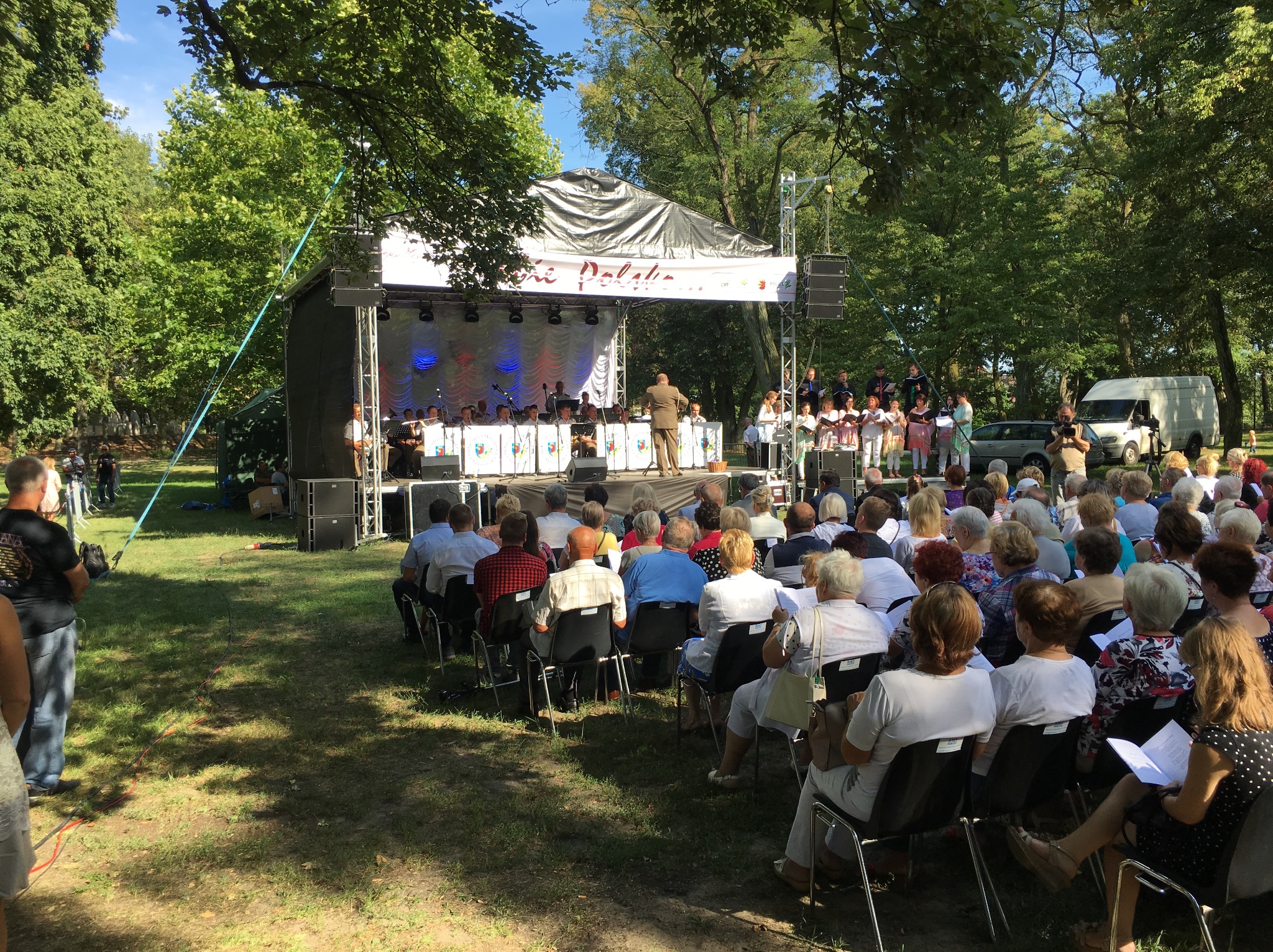 Plenerowy koncert chóru i orkiestry na scenie z białym tłem i srebrną konstrukcją zadaszenia, widownia siedząca na krzesłach na trawie, w tle drzewa i biały bus.