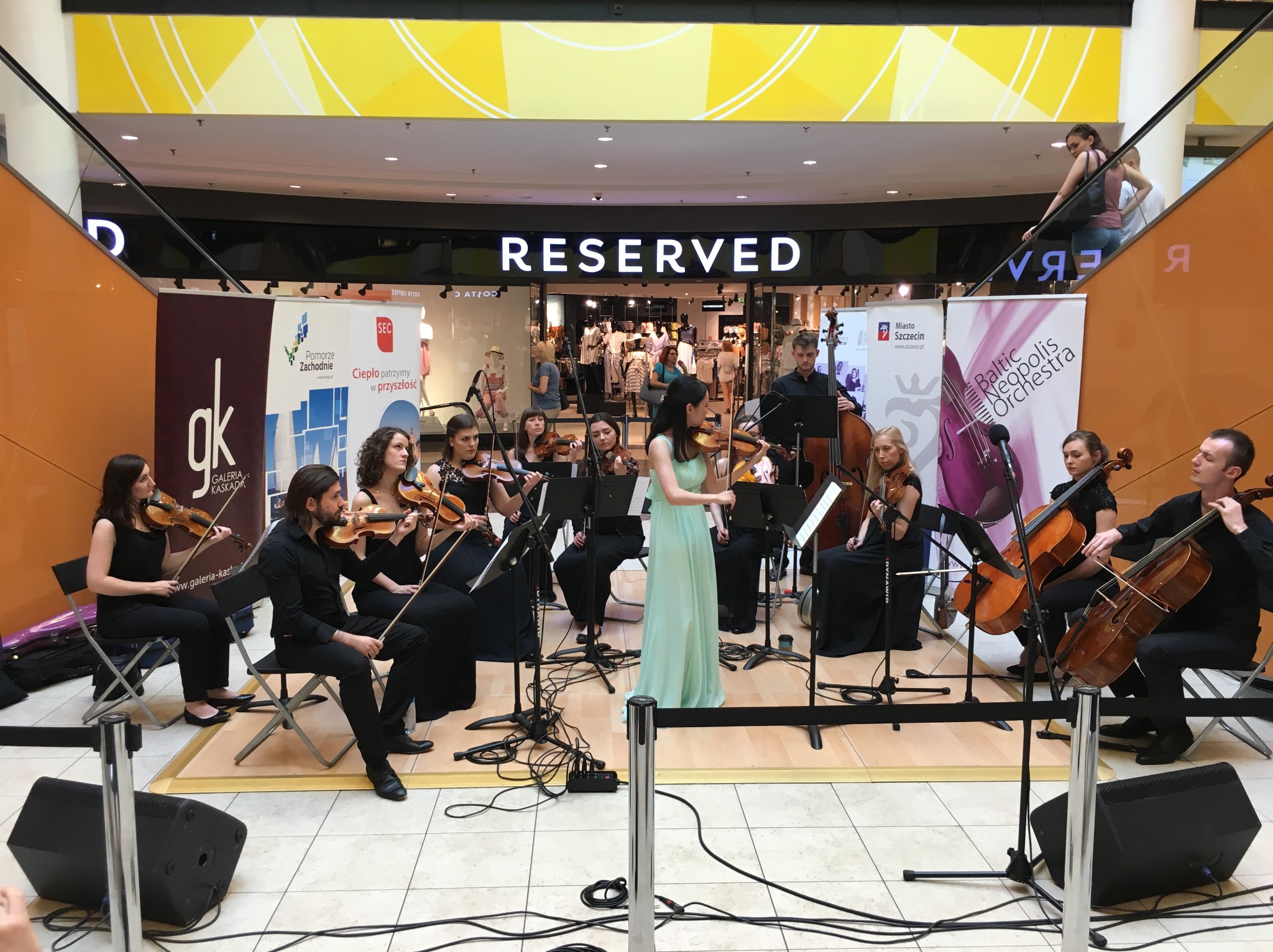Kameralny występ orkiestry smyczkowej w centrum handlowym przed sklepem Reserved, widoczne skrzypce, wiolonczele, kontrabas, czarne kolumny głośnikowe, plakat z logo orkiestry.