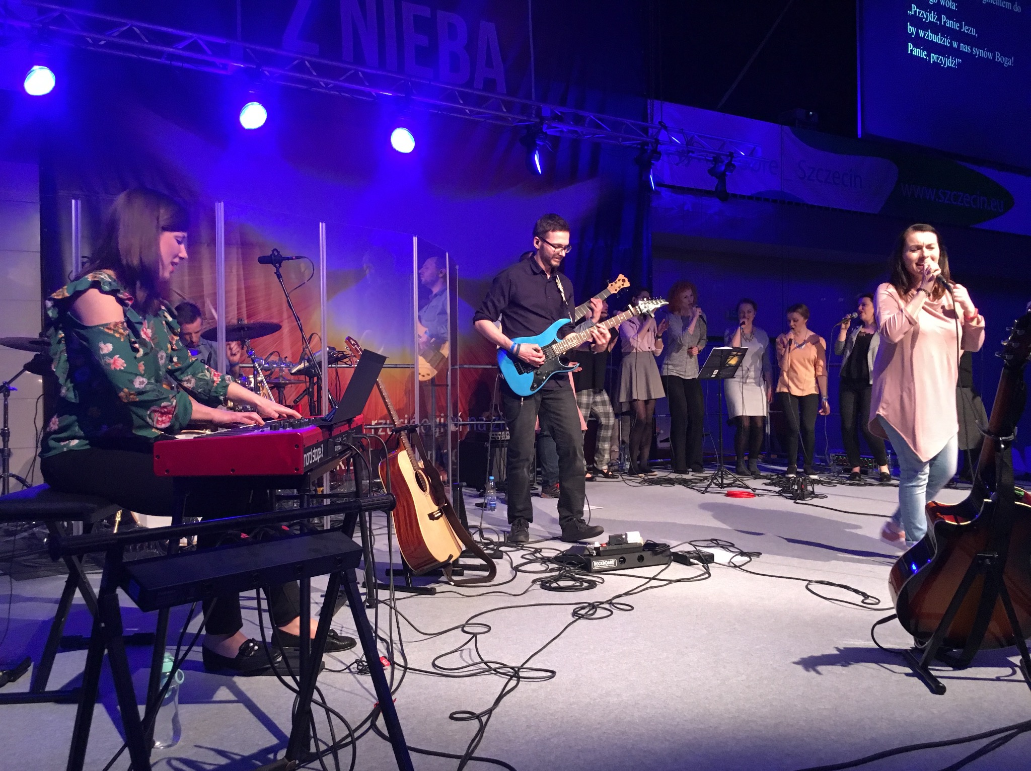 Zespół muzyczny na scenie, wokalistka z mikrofonem, gitarzysta z niebieską gitarą elektryczną, klawiszowiec z czerwonym keyboardem, chórki w tle, scena oświetlona niebieskim światłem, widoczne...