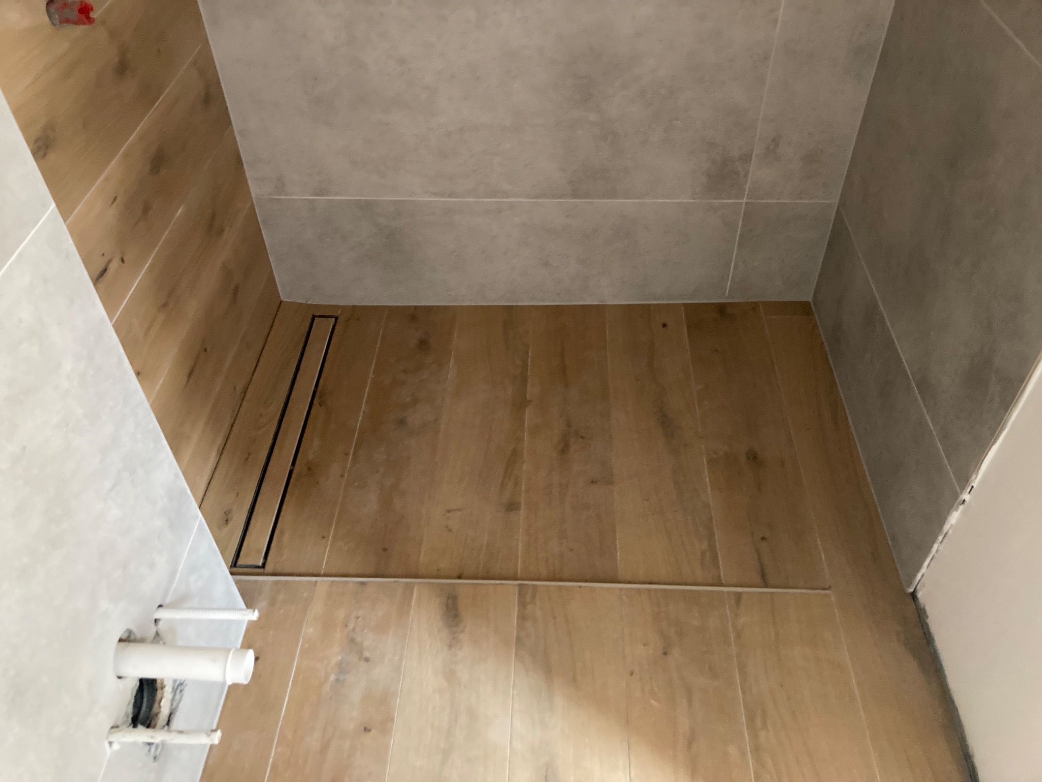 Wykończona kabina prysznicowa z drewnopodobnymi płytkami na podłodze i ścianach, z odpływem liniowym i widocznymi rurami instalacji wodnej.