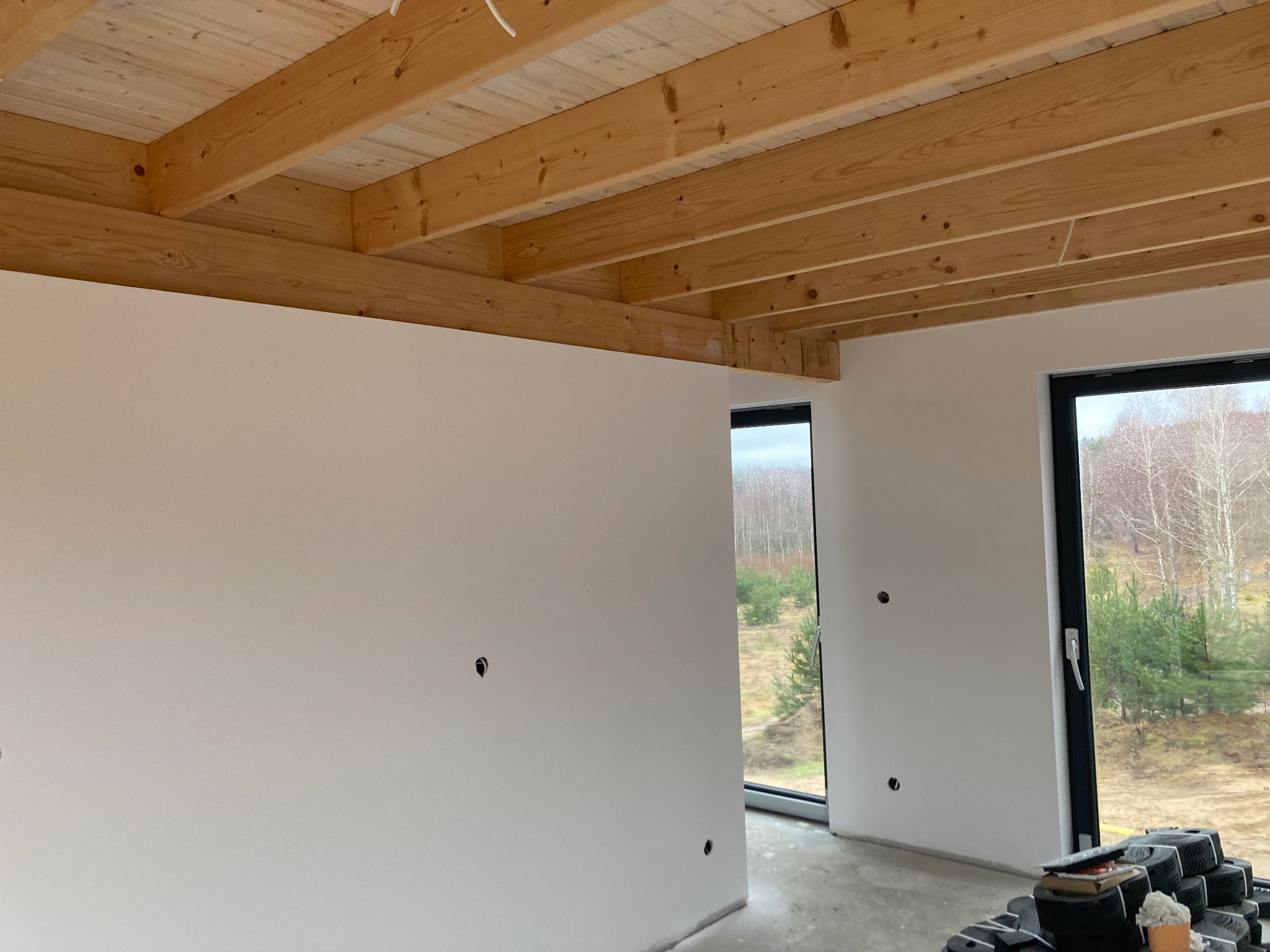 Wnętrze w trakcie wykańczania: gładkie, białe ściany z widocznymi puszkami elektrycznymi, surowy drewniany sufit z belek, okna z widokiem na zewnątrz, na podłodze leżą materiały budowlane.