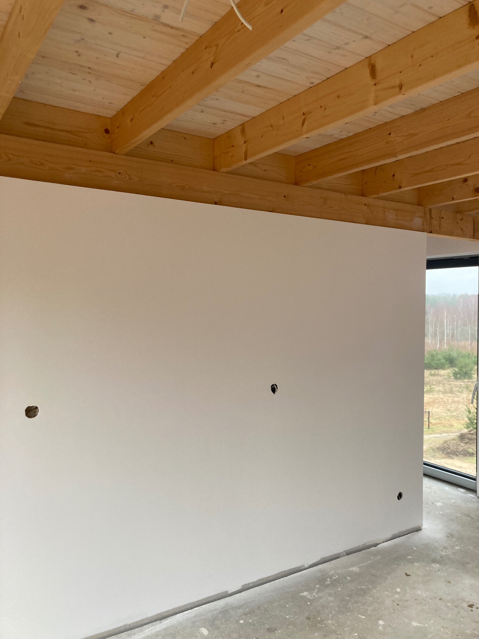 Wnętrze w trakcie prac wykończeniowych: gładka, biała ściana z widocznymi otworami na gniazdka i surowy beton na podłodze, drewniany strop z belek, okno z widokiem na las w tle.