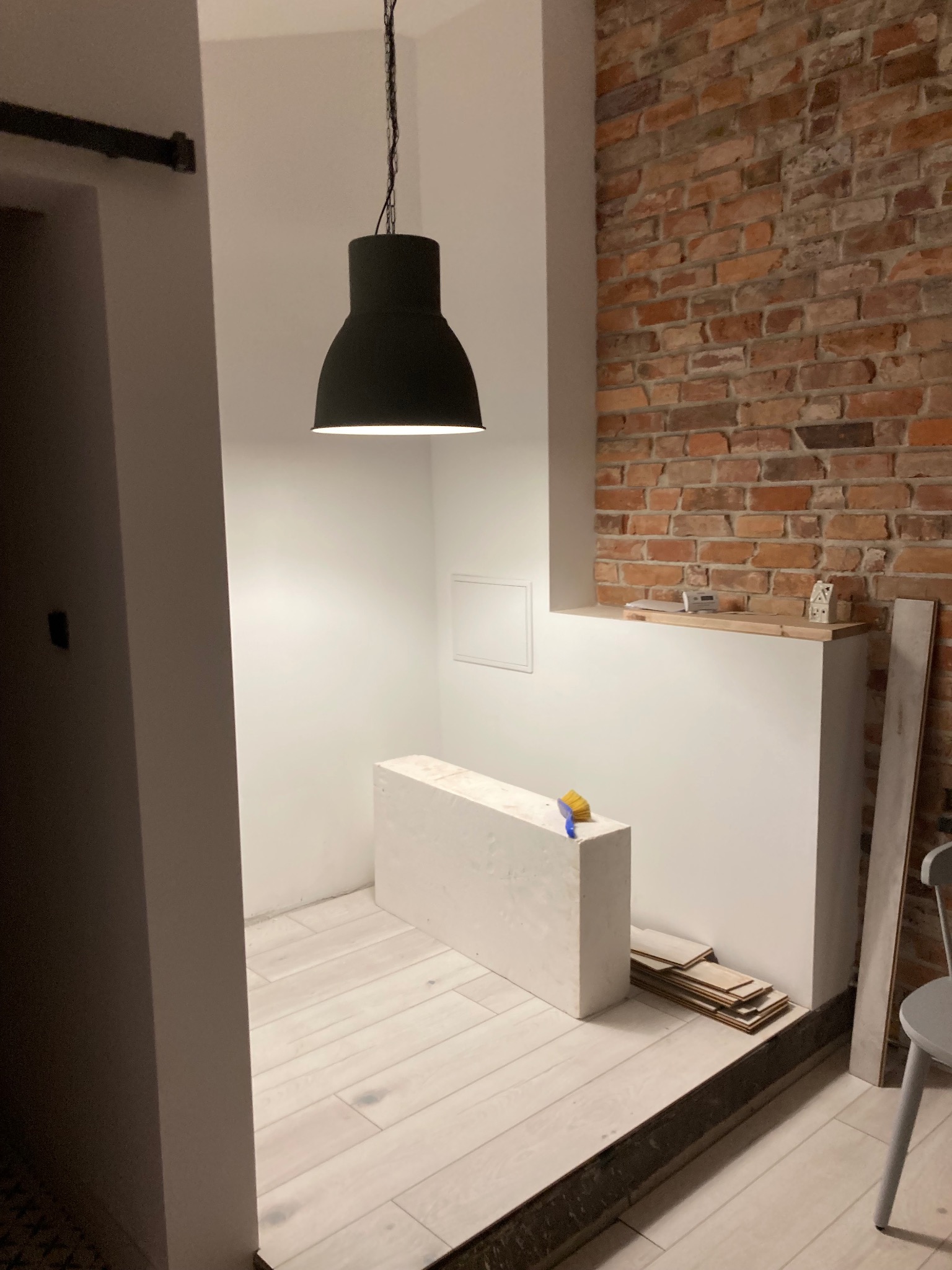 Wnętrze w trakcie remontu: świeżo ułożona jasna drewniana podłoga, fragment ściany z cegły, wisząca czarna lampa, przygotowane panele podłogowe, szczotka na białym podwyższeniu.