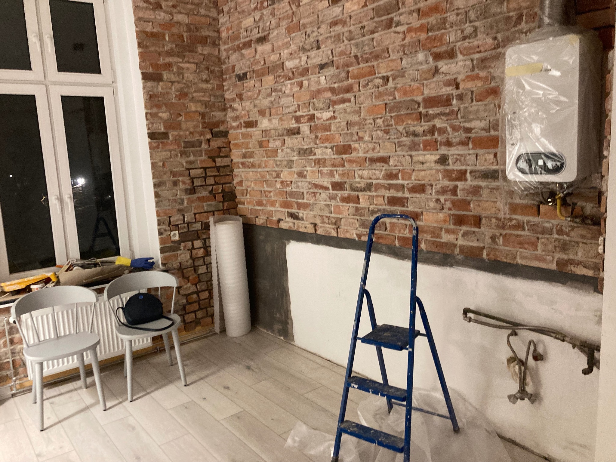 Wnętrze w trakcie odświeżania: surowa ceglana ściana kontrastuje z fragmentem pomalowanej ściany, niebieska drabina, dwa krzesła z torbą, nowy piec gazowy w folii ochronnej.