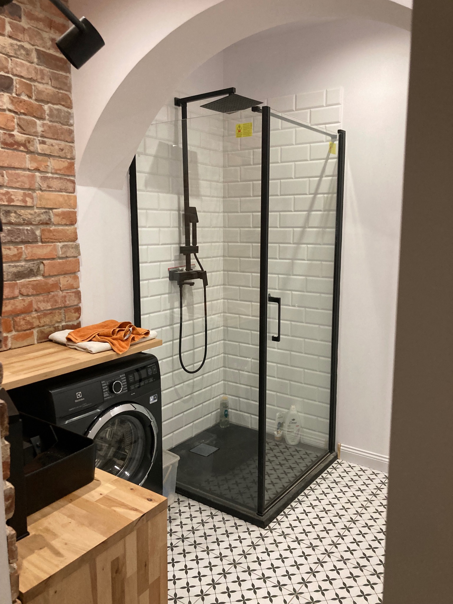 Nowoczesna łazienka z kabiną prysznicową typu walk-in z czarnymi profilami, białe płytki imitujące cegłę, pralka Electrolux z drewnianym blatem i pomarańczowym ręcznikiem, podłoga w biało-czarne...