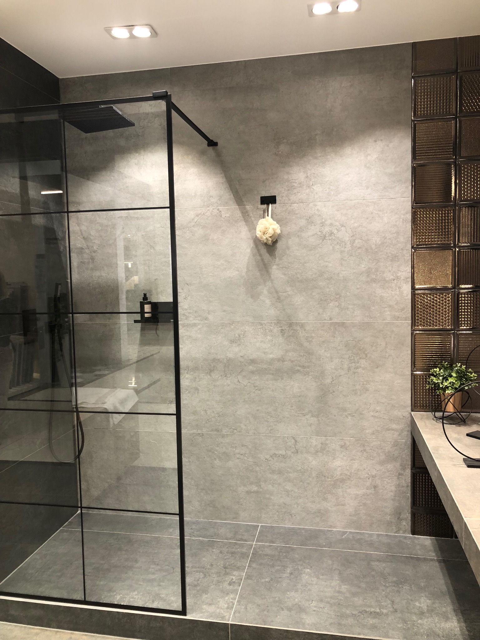 Nowoczesna łazienka z szarą, matową okładziną ścienną, kabiną prysznicową typu walk-in z czarnymi profilami, dekoracyjną ścianką z luksferów i minimalistyczną umywalką z rośliną doniczkową.