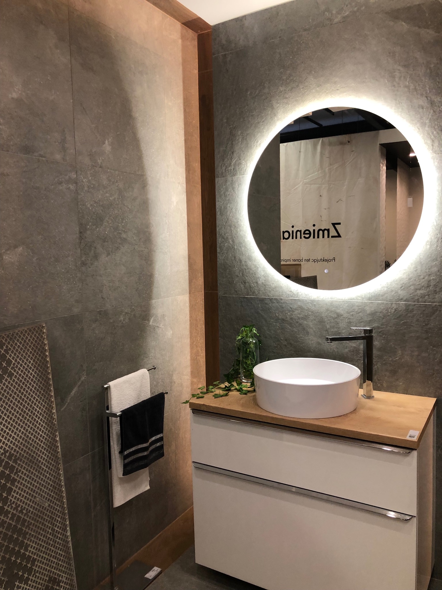 Nowoczesna łazienka w Oostzaan z umywalką nablatową, baterią i okrągłym lustrem z podświetleniem LED na tle szarych płytek imitujących kamień.