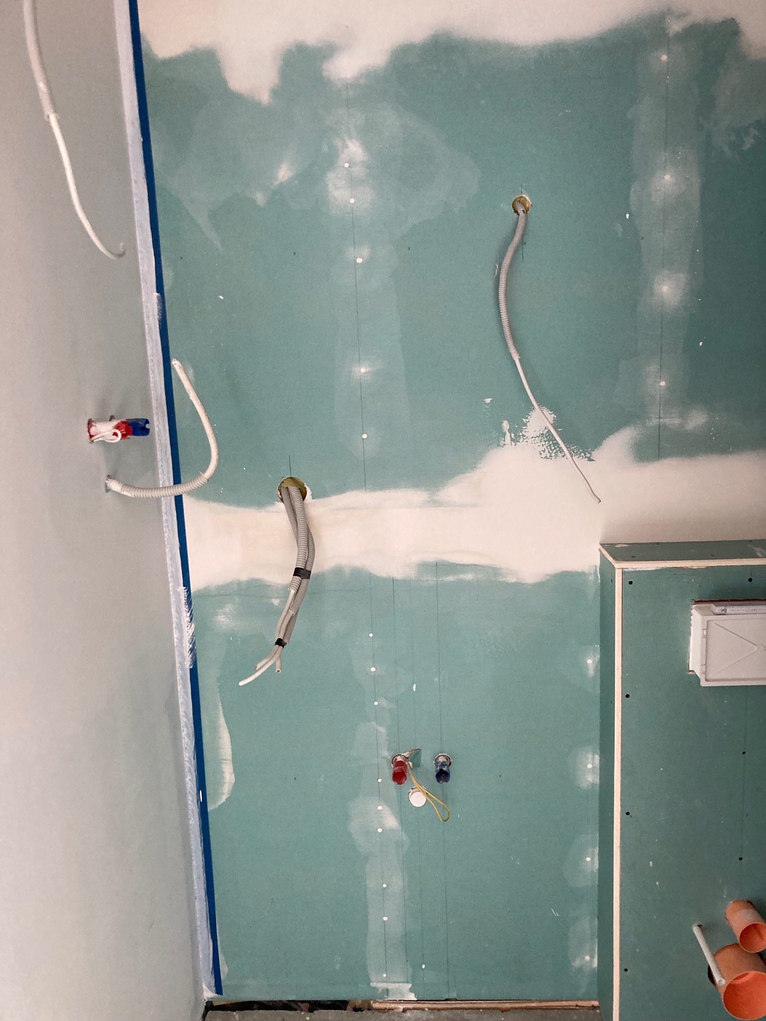 Ściana w trakcie przygotowania do wykończenia, płyty gipsowo-kartonowe z widocznymi przewodami elektrycznymi i rurami hydraulicznymi, szpachlowane łączenia płyt.
