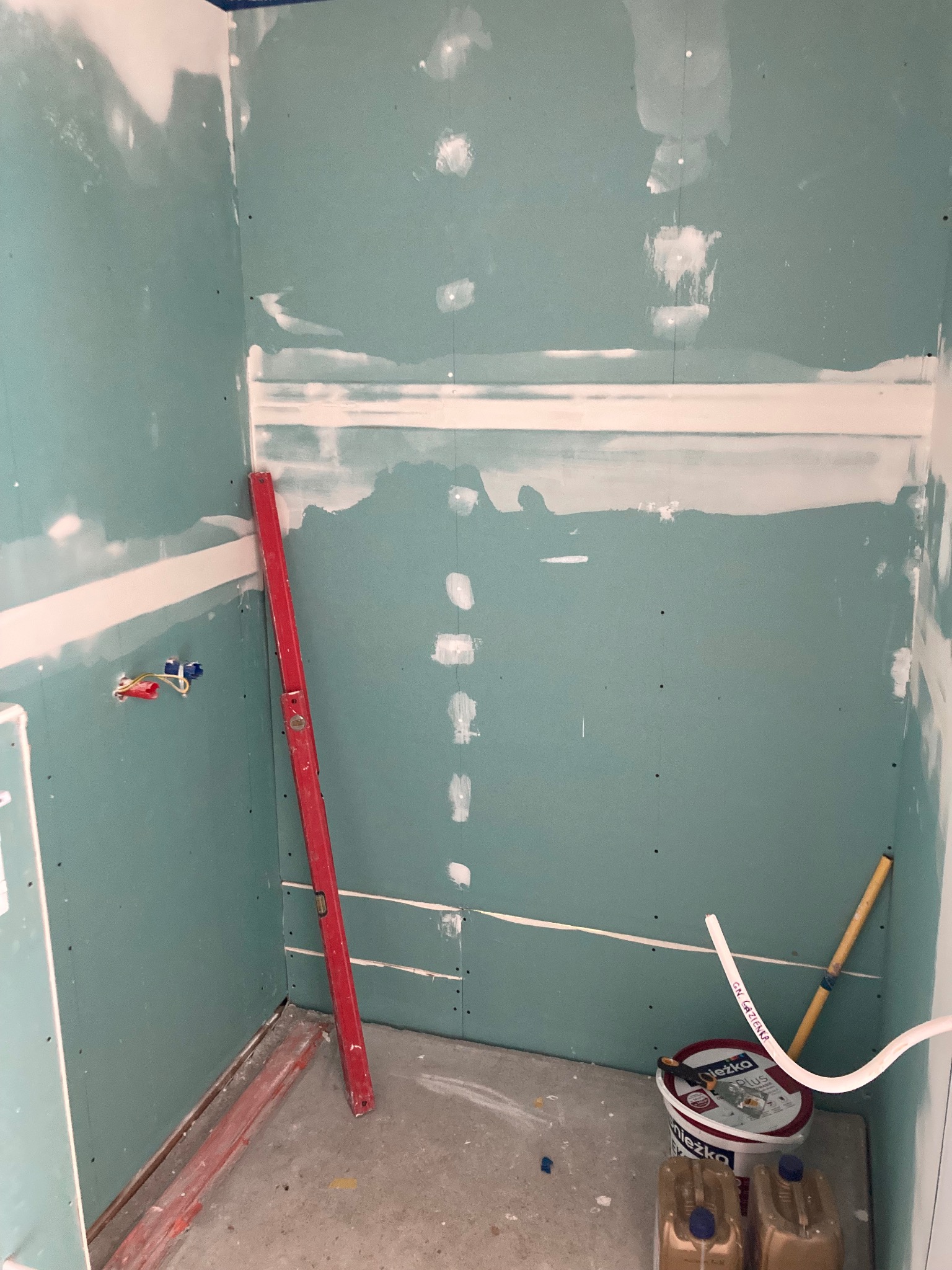 Wnętrze łazienki w trakcie prac wykończeniowych: ściany obłożone płytami gipsowo-kartonowymi, częściowo zaszpachlowane, widoczna poziomica oparta o ścianę, wiadro z masą szpachlową i kanistry...