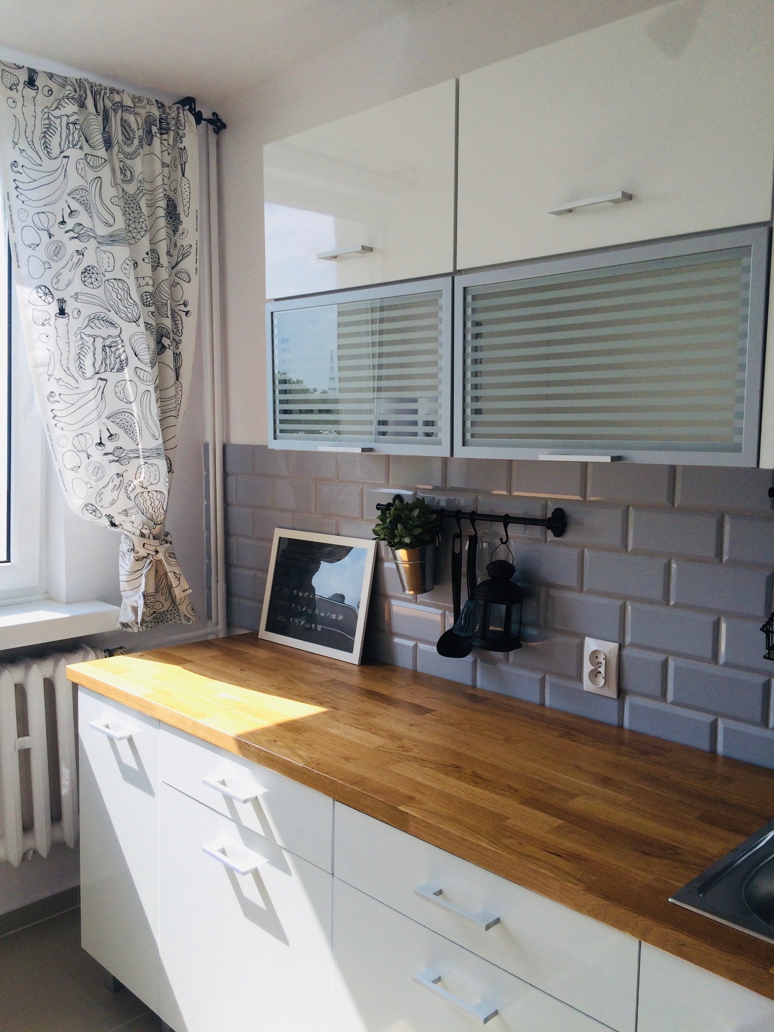 Jasna kuchnia z drewnianym blatem, szarymi płytkami metra i białymi szafkami, okno z zasłoną w warzywne wzory, dekoracje na ścianie.