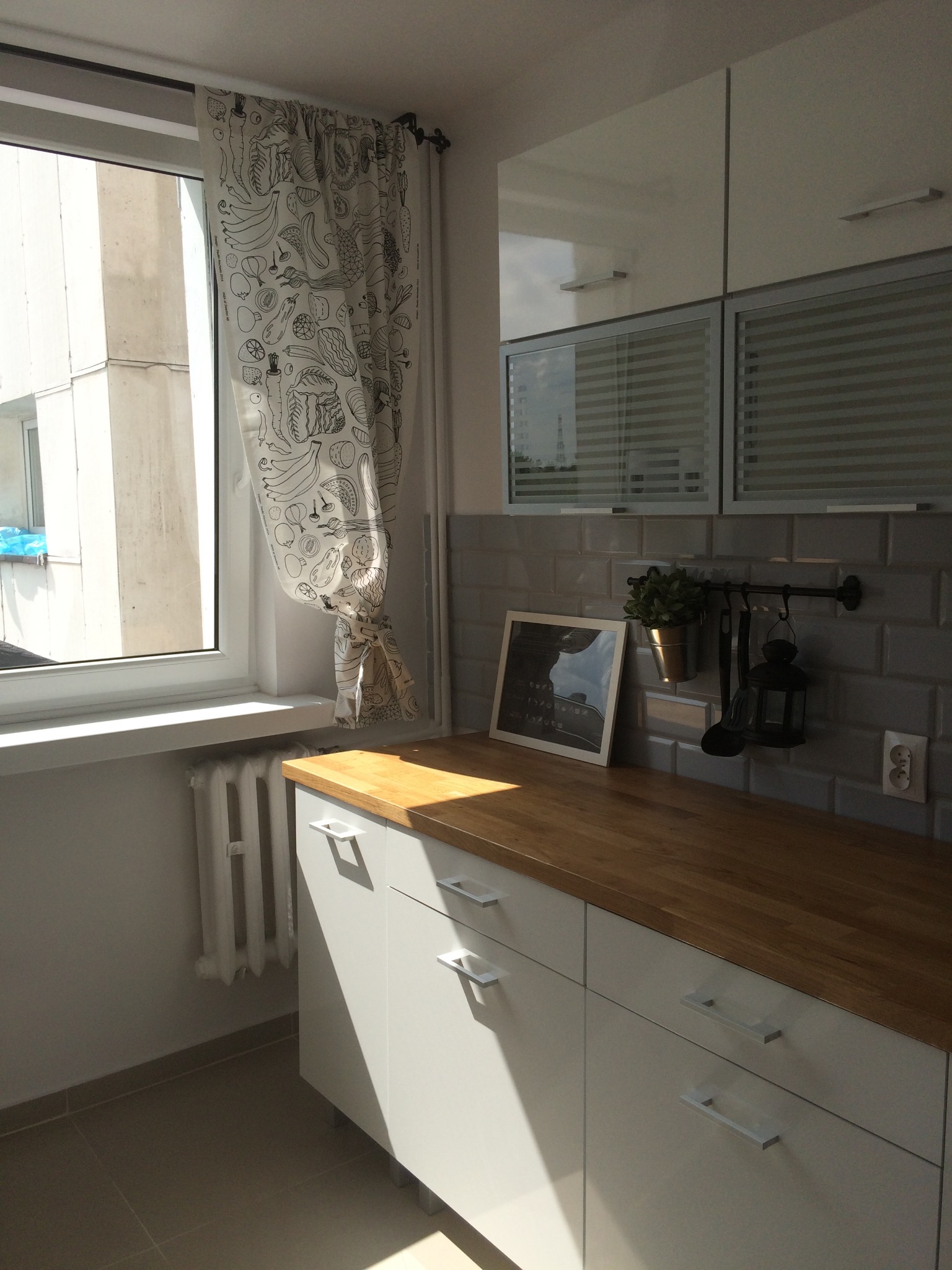 Jasna kuchnia z drewnianym blatem, szarymi kafelkami na ścianie, białymi szafkami i zasłoną w oknie z motywem warzyw i owoców.