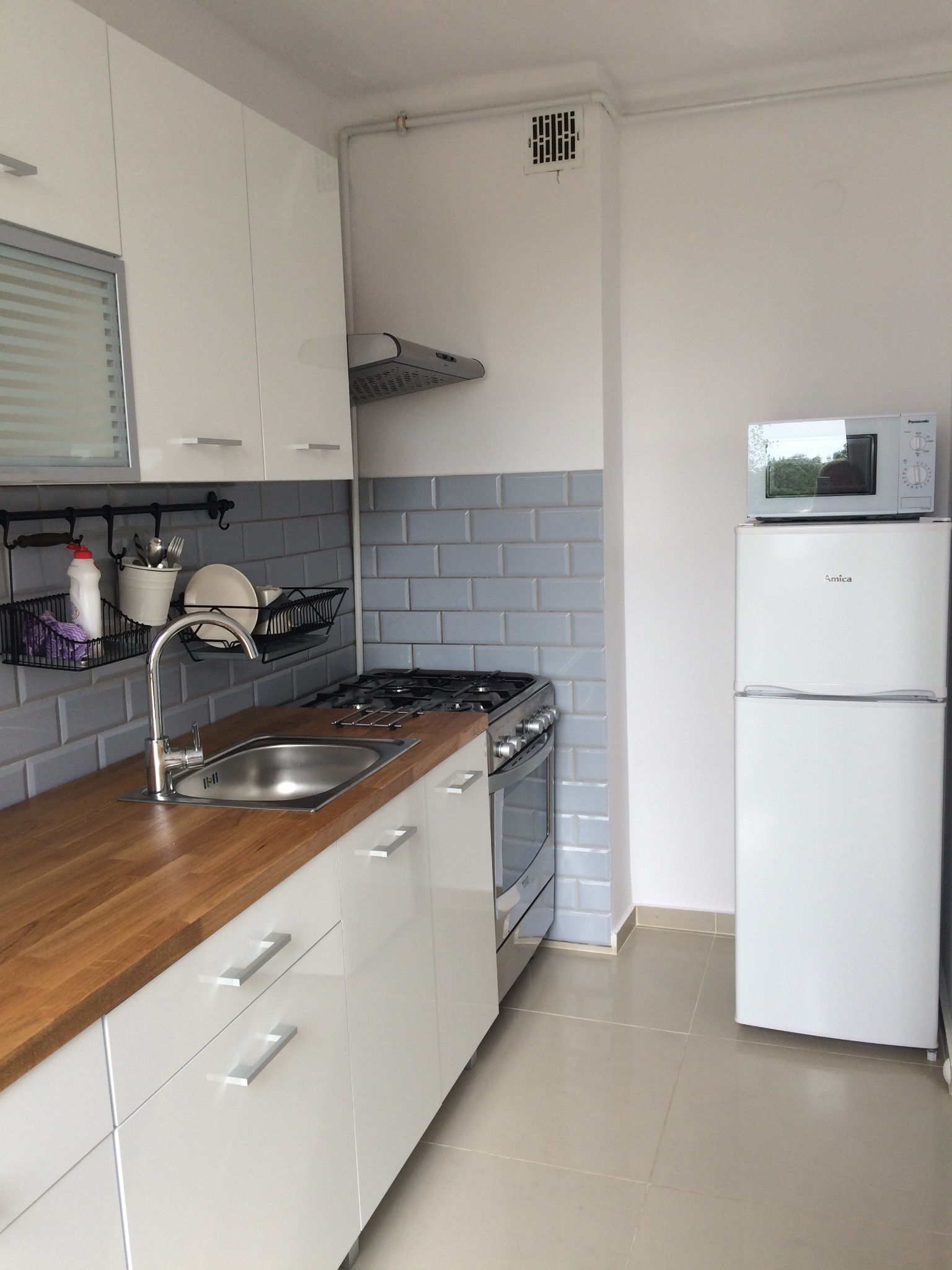 Nowoczesna, jasna kuchnia z drewnianym blatem, szarymi płytkami ściennymi i białymi meblami, uzupełniona o kuchenkę gazową, srebrny zlew i białą lodówkę z mikrofalówką na górze.