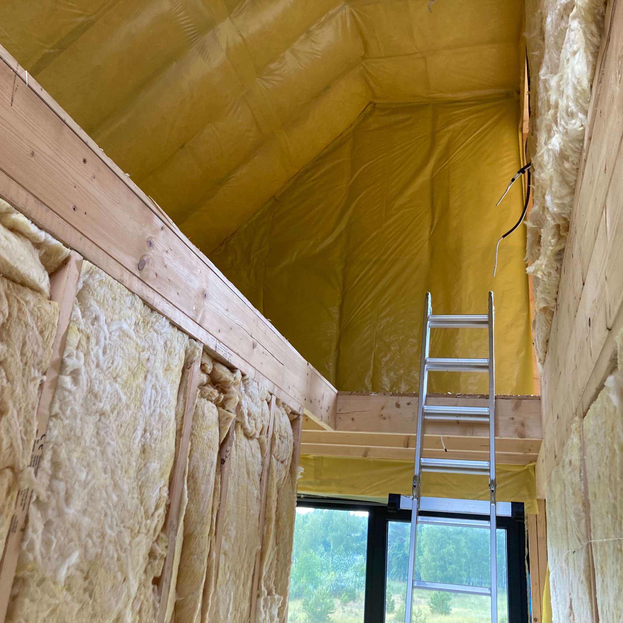 Ocieplenie poddasza wełną mineralną z folią paroizolacyjną, widoczna drabina aluminiowa i konstrukcja drewniana, okno z widokiem na zieleń na zewnątrz.