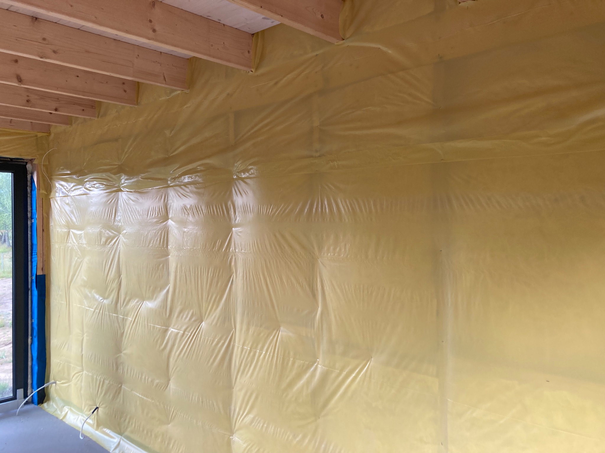 Ściana wewnętrzna zabezpieczona żółtą folią budowlaną przed dalszymi pracami, widoczne drewniane elementy konstrukcyjne sufitu oraz kable elektryczne wystające z podłogi przy ościeżnicy okna.