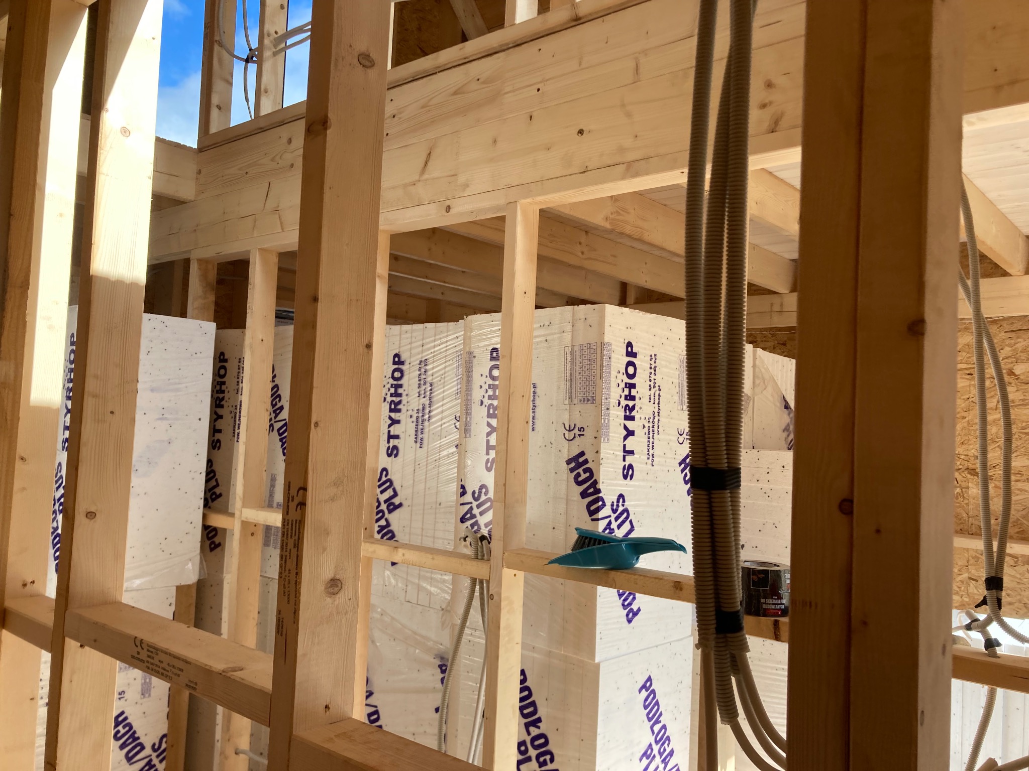 Wnętrze domu w trakcie budowy, widoczne drewniane ramy konstrukcyjne, izolacja ze styropianu z folią, przewody elektryczne i niebieska zmiotka na drewnianej belce.