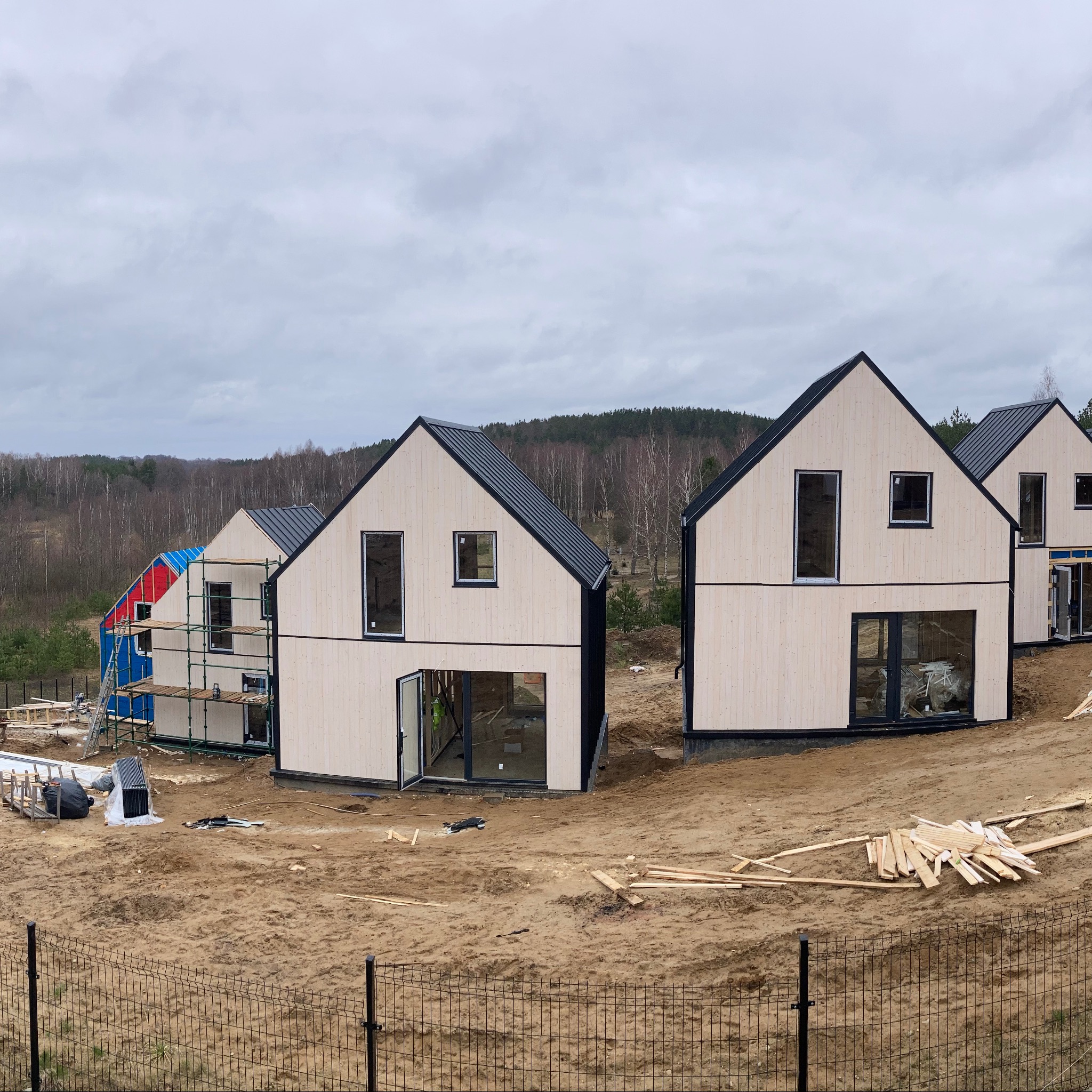 Wznoszone domy modułowe z jasną elewacją drewnianą i czarnymi ramami okiennymi, jeden w trakcie montażu z rusztowaniem i folią ochronną na dachu, otoczone piaszczystym terenem budowy.