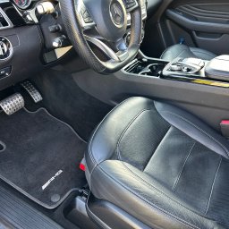 AS-TJ SPÓŁKA Z OGRANICZONĄ ODPOWIEDZIALNOŚCIĄ - Wnętrze samochodu Mercedes-Benz AMG z czarną skórzaną tapicerką, widoczny fotel kierowcy, dywaniki z logo AMG i elementy deski rozdzielczej.