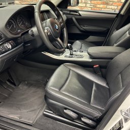 AS-TJ SPÓŁKA Z OGRANICZONĄ ODPOWIEDZIALNOŚCIĄ - Wnętrze samochodu BMW X Line z czarną skórzaną tapicerką, widok na fotel kierowcy, deskę rozdzielczą i fragment ceglanej ściany w tle.