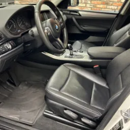 Wnętrze samochodu BMW X Line z czarną skórzaną tapicerką, widok na fotel kierowcy, deskę rozdzielczą i fragment ceglanej ściany w tle.