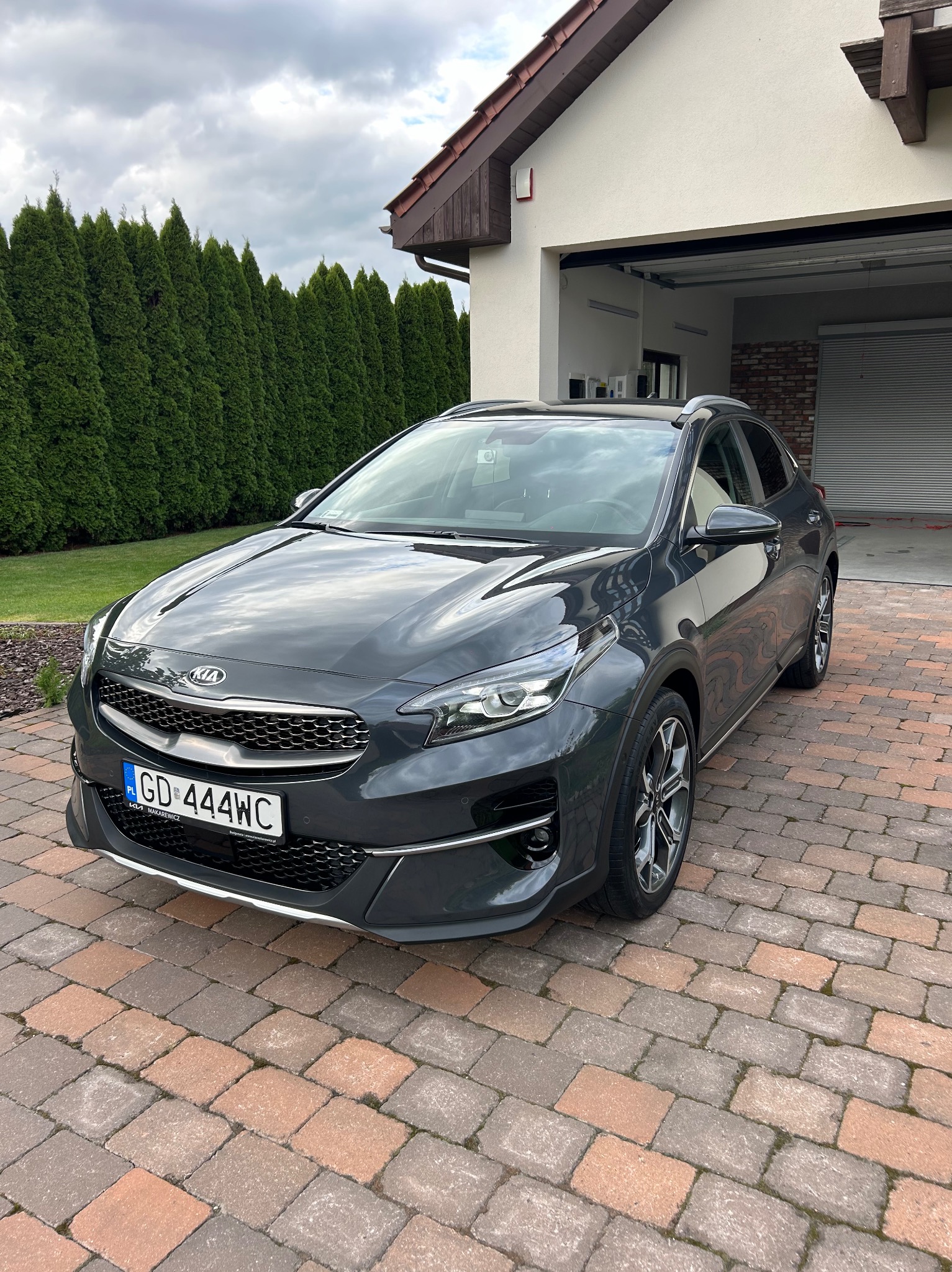 Szary samochód Kia XCeed zaparkowany na kostce brukowej przed garażem i żywopłotem z tui.