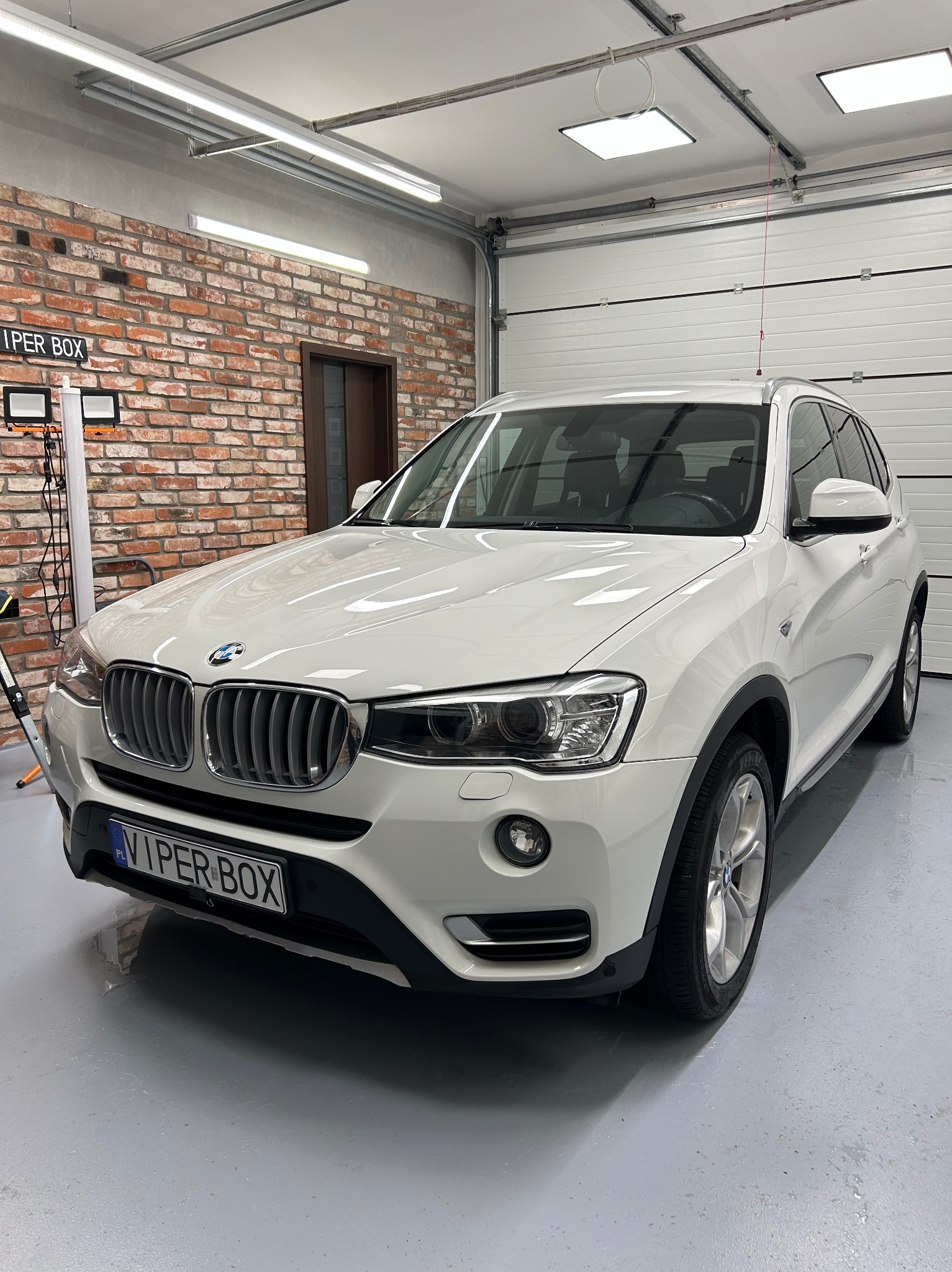 Białe BMW X3 po umyciu, stojące w garażu z ceglaną ścianą i bramą segmentową, widoczny połysk na karoserii i mokra posadzka.