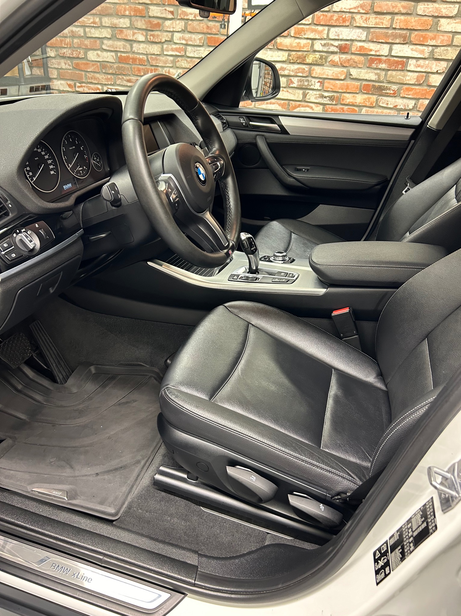 Wnętrze samochodu BMW X Line z czarną skórzaną tapicerką, widok na fotel kierowcy, deskę rozdzielczą i fragment ceglanej ściany w tle.