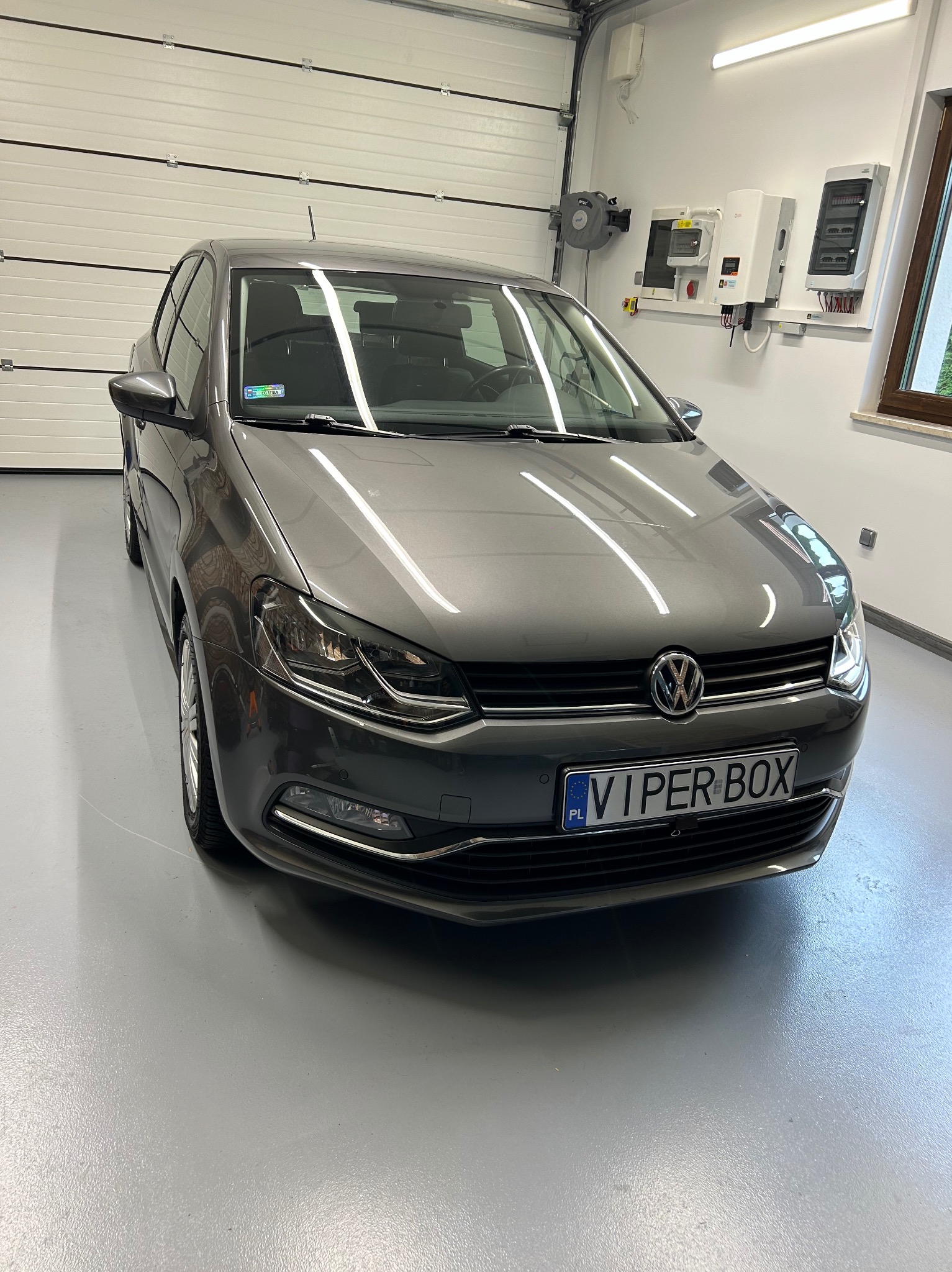 Szary Volkswagen Polo po umyciu, stojący w czystym garażu z białą bramą segmentową i instalacją elektryczną na ścianie.