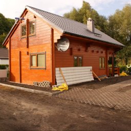 Akka II s.c. Kanty Waldemar, Kanty Daniela - Nowy, drewniany domek z brązowymi ramami okiennymi i anteną satelitarną na ścianie, częściowo wykończony teren wokół z ażurowymi płytami.