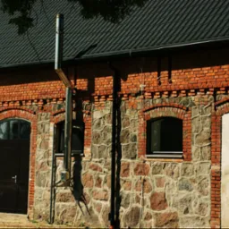 Murowany budynek z kamienno-ceglanymi ścianami, czarną stolarką okienną i drzwiową, oraz zewnętrznym kominem ze stali nierdzewnej.