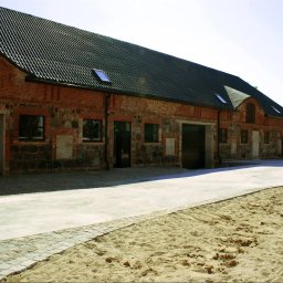 Akka II s.c. Kanty Waldemar, Kanty Daniela - Duży, odnowiony budynek gospodarczy z czerwonej cegły i kamienia, z nowymi drzwiami garażowymi i oknami, z pracownikiem w niebieskiej koszuli w paski w otwartych drzwiach. Nowy betonowy chodnik...