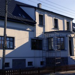 Akka II s.c. Kanty Waldemar, Kanty Daniela - Dwupiętrowy dom z beżową elewacją, ciemną dachówką, brązowymi ramami okiennymi i metalowym rusztowaniem przy elewacji frontowej.