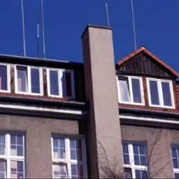 Elewacja budynku z białymi oknami, czerwonym dachem z dachówki i szarym kominem na tle błękitnego nieba, widoczne anteny na dachu.