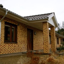 Akka II s.c. Kanty Waldemar, Kanty Daniela - Dom jednorodzinny z żółtej cegły z brązowymi oknami i drzwiami, widok z zewnątrz w pochmurny dzień, z widoczną roślinnością i elementami otoczenia.