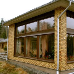 Akka II s.c. Kanty Waldemar, Kanty Daniela - Duże okno z brązowymi ramami w domu z żółtej cegły, widok z zewnątrz, widoczne drzewa odbijające się w szybach, zasłony w oknach.