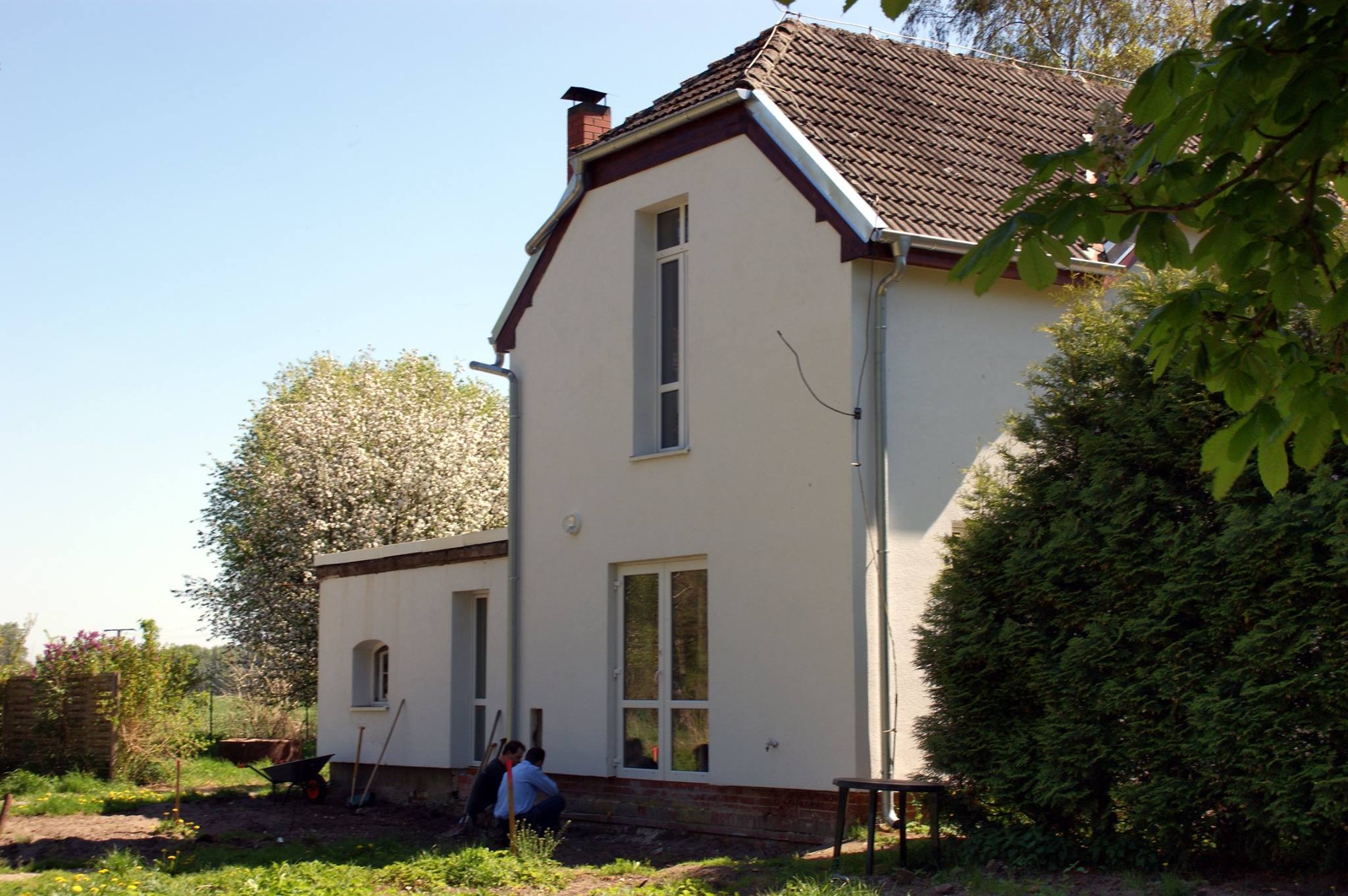 Boczny widok domu z nowymi oknami i drzwiami, dwóch pracowników siedzi na ziemi podczas przerwy, na trawniku leżą narzędzia, a w tle kwitnące drzewo.