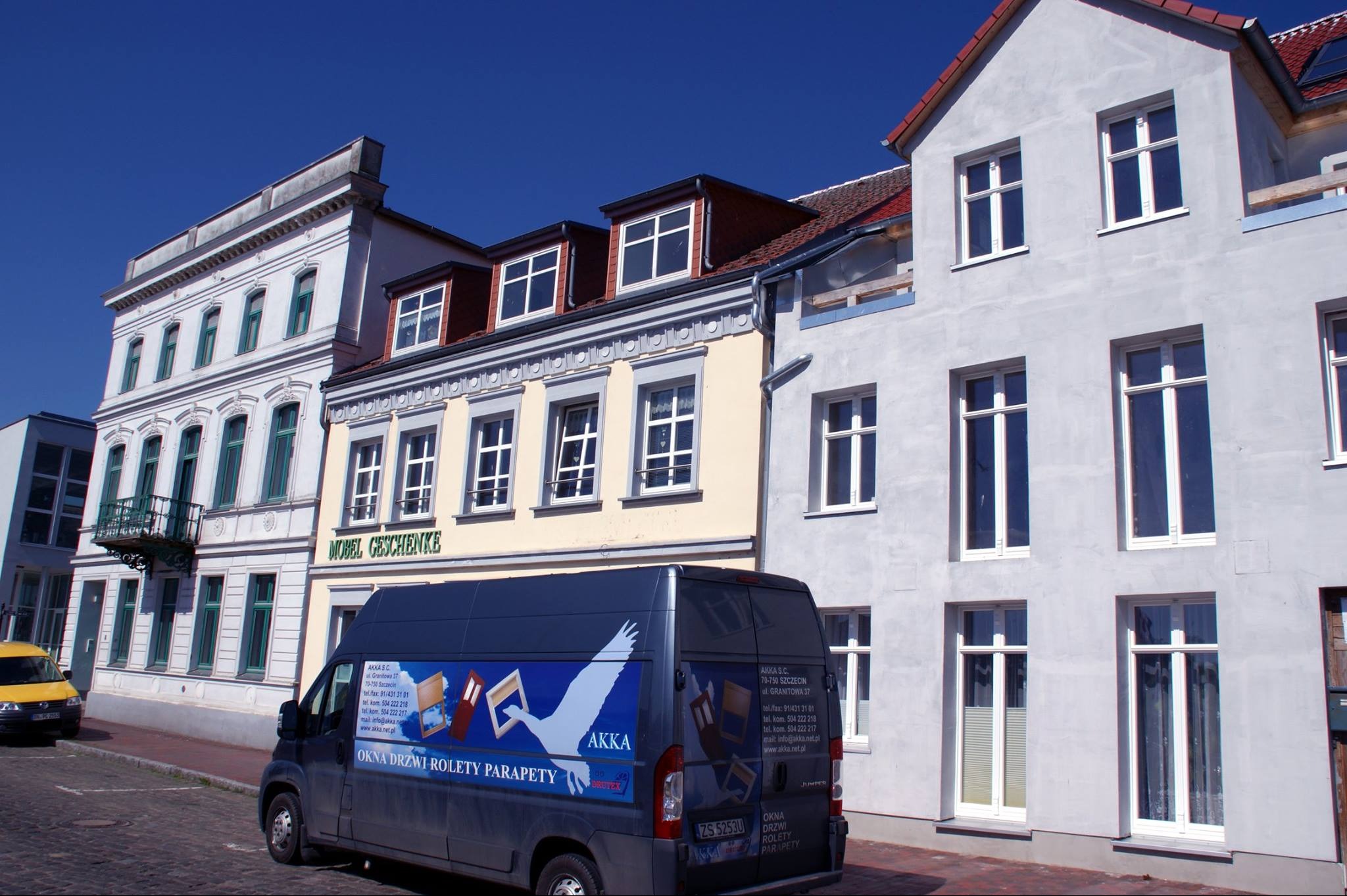 Ulica z trzema kamienicami o różnej architekturze, na pierwszym planie zaparkowany samochód dostawczy z reklamą okien, drzwi i rolet firmy AKKA.