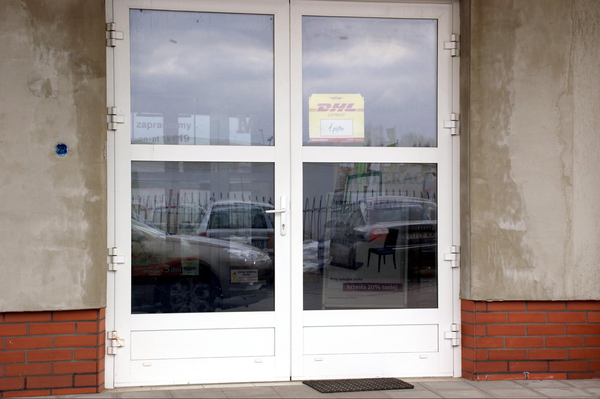 Białe, dwuskrzydłowe drzwi wejściowe z PCV do lokalu, z naklejonymi reklamami i informacjami, widoczne odbicia samochodów i wnętrza sklepu.