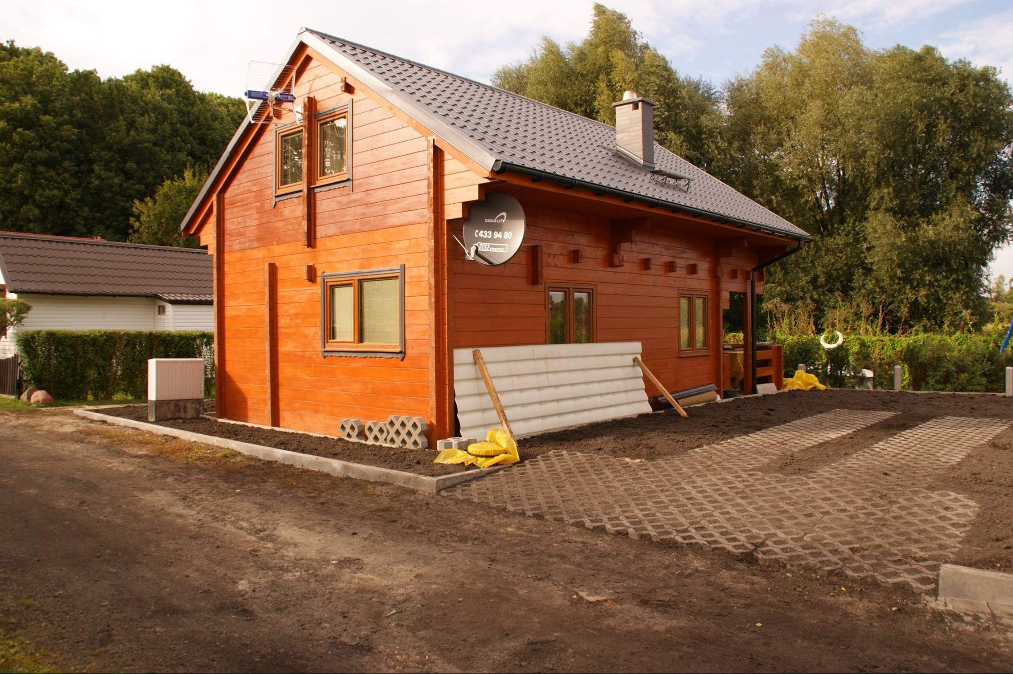 Nowy, drewniany domek z brązowymi ramami okiennymi i anteną satelitarną na ścianie, częściowo wykończony teren wokół z ażurowymi płytami.
