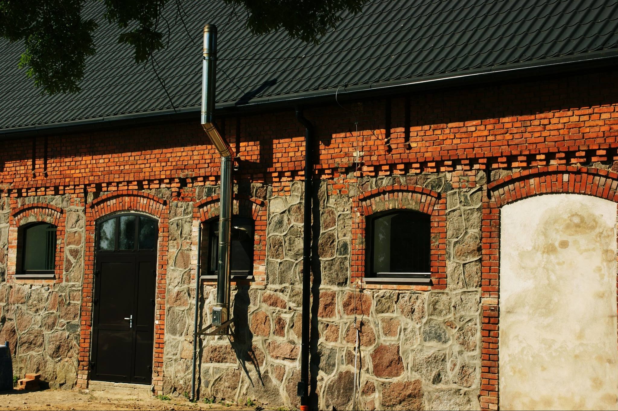 Murowany budynek z kamienno-ceglanymi ścianami, czarną stolarką okienną i drzwiową, oraz zewnętrznym kominem ze stali nierdzewnej.