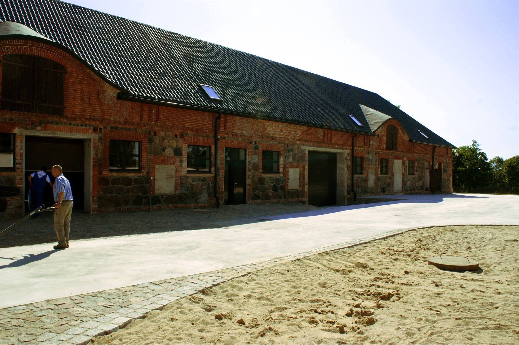 Duży, odnowiony budynek gospodarczy z czerwonej cegły i kamienia, z nowymi drzwiami garażowymi i oknami, z pracownikiem w niebieskiej koszuli w paski w otwartych drzwiach. Nowy betonowy chodnik...