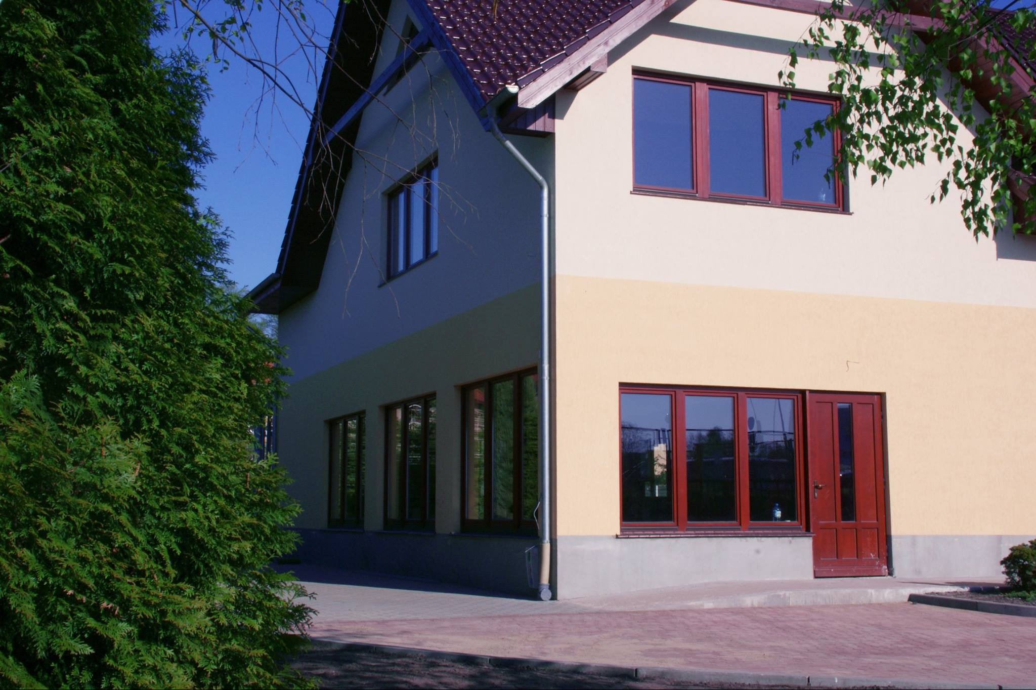 Dwukondygnacyjny dom z beżową elewacją i brązowymi ramami okiennymi oraz drzwiami wejściowymi, widoczny z zewnątrz, z fragmentem zielonego drzewa iglastego po lewej stronie.