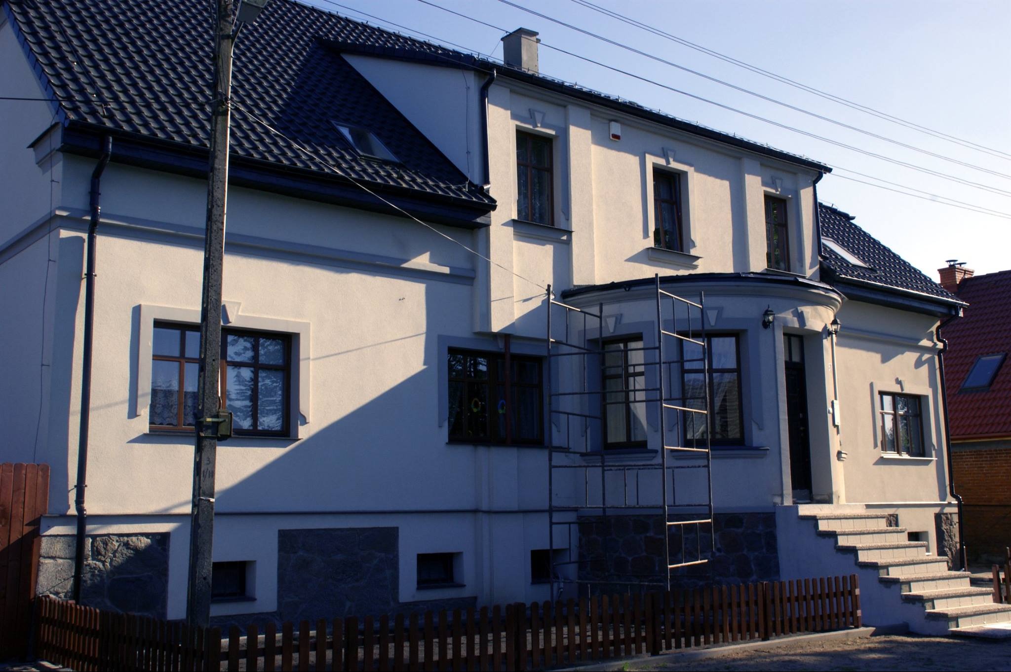 Dwupiętrowy dom z beżową elewacją, ciemną dachówką, brązowymi ramami okiennymi i metalowym rusztowaniem przy elewacji frontowej.