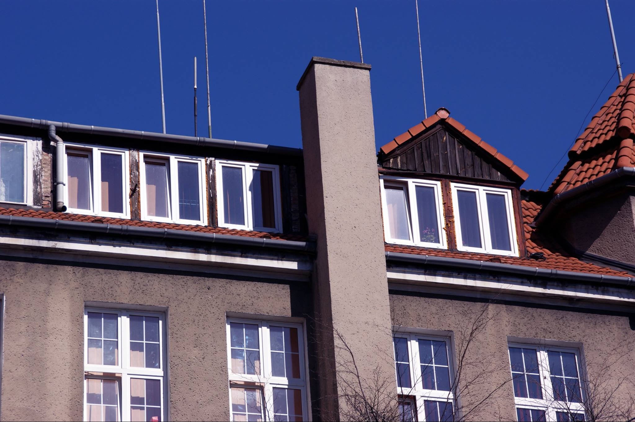 Elewacja budynku z białymi oknami, czerwonym dachem z dachówki i szarym kominem na tle błękitnego nieba, widoczne anteny na dachu.