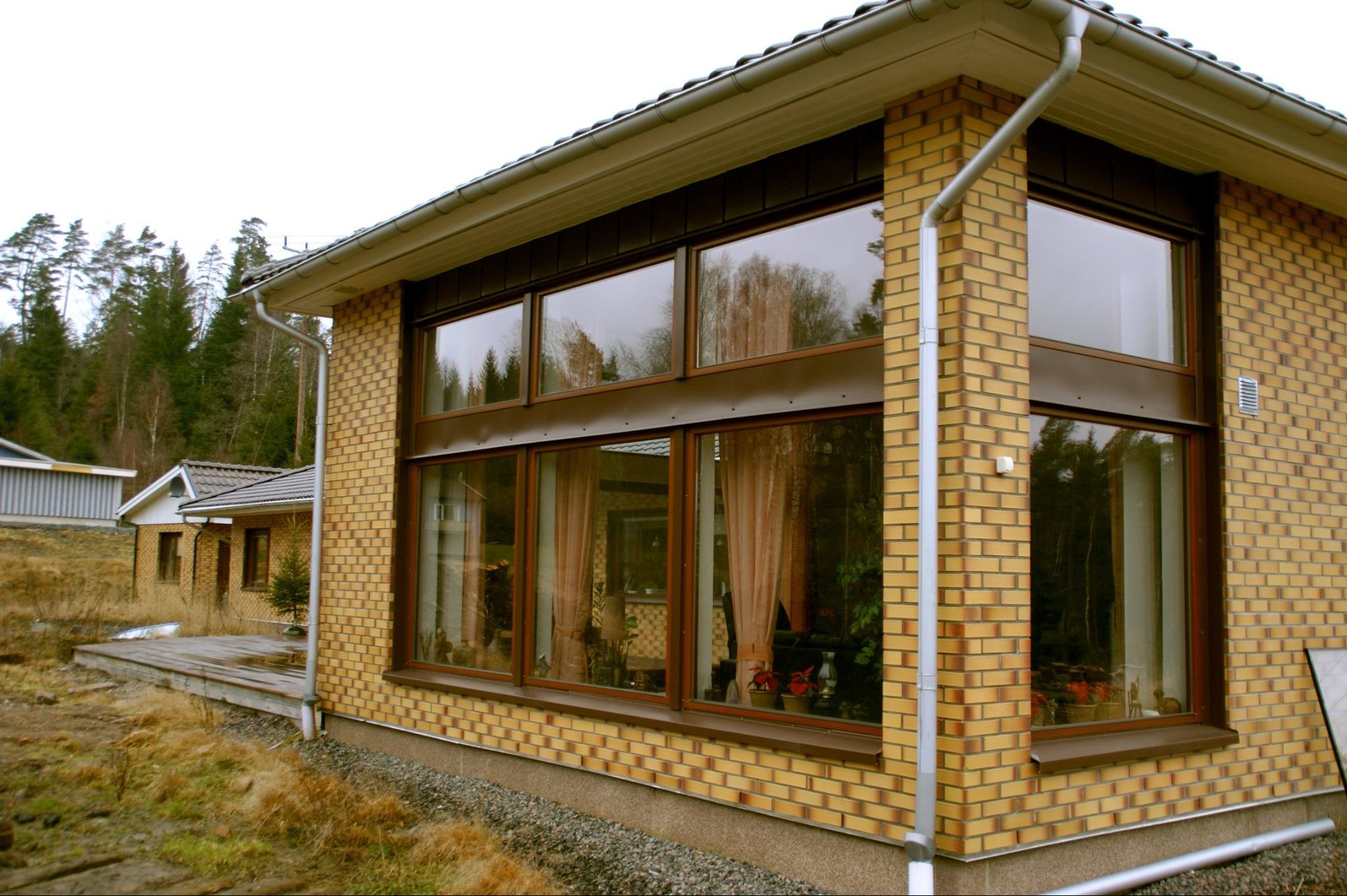 Duże okno z brązowymi ramami w domu z żółtej cegły, widok z zewnątrz, widoczne drzewa odbijające się w szybach, zasłony w oknach.