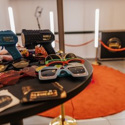 Klemczak FotoShow - Akcesoria do sesji zdjęciowej: pistolety na bańki mydlane, okulary w różnych kształtach i rekwizyty leżące na okrągłym stole, w tle widoczny automat do zdjęć z czerwonym sznurem.