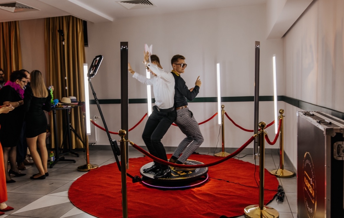 Dwaj mężczyźni w zabawnych pozach na obrotowej platformie, otoczonej czerwonym dywanem i elementami dekoracyjnymi, podczas sesji zdjęciowej z wykorzystaniem lamp i akcesoriów.